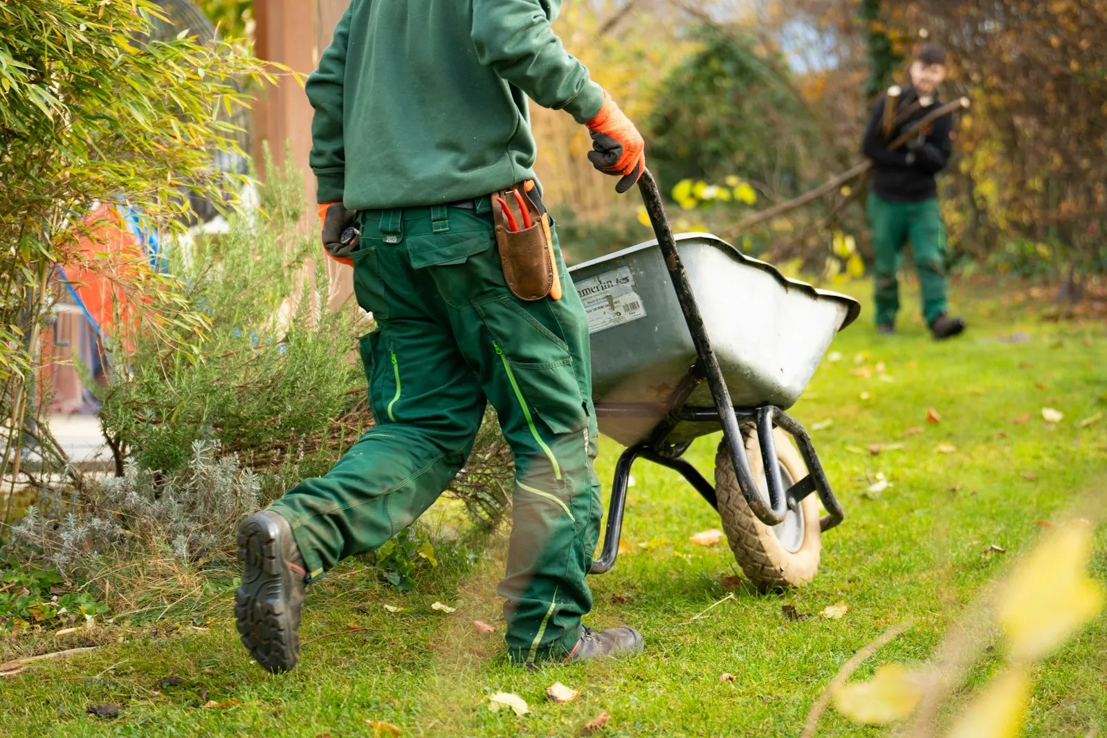 A crew member in green work clothes wheeling a barrow across a lawn
