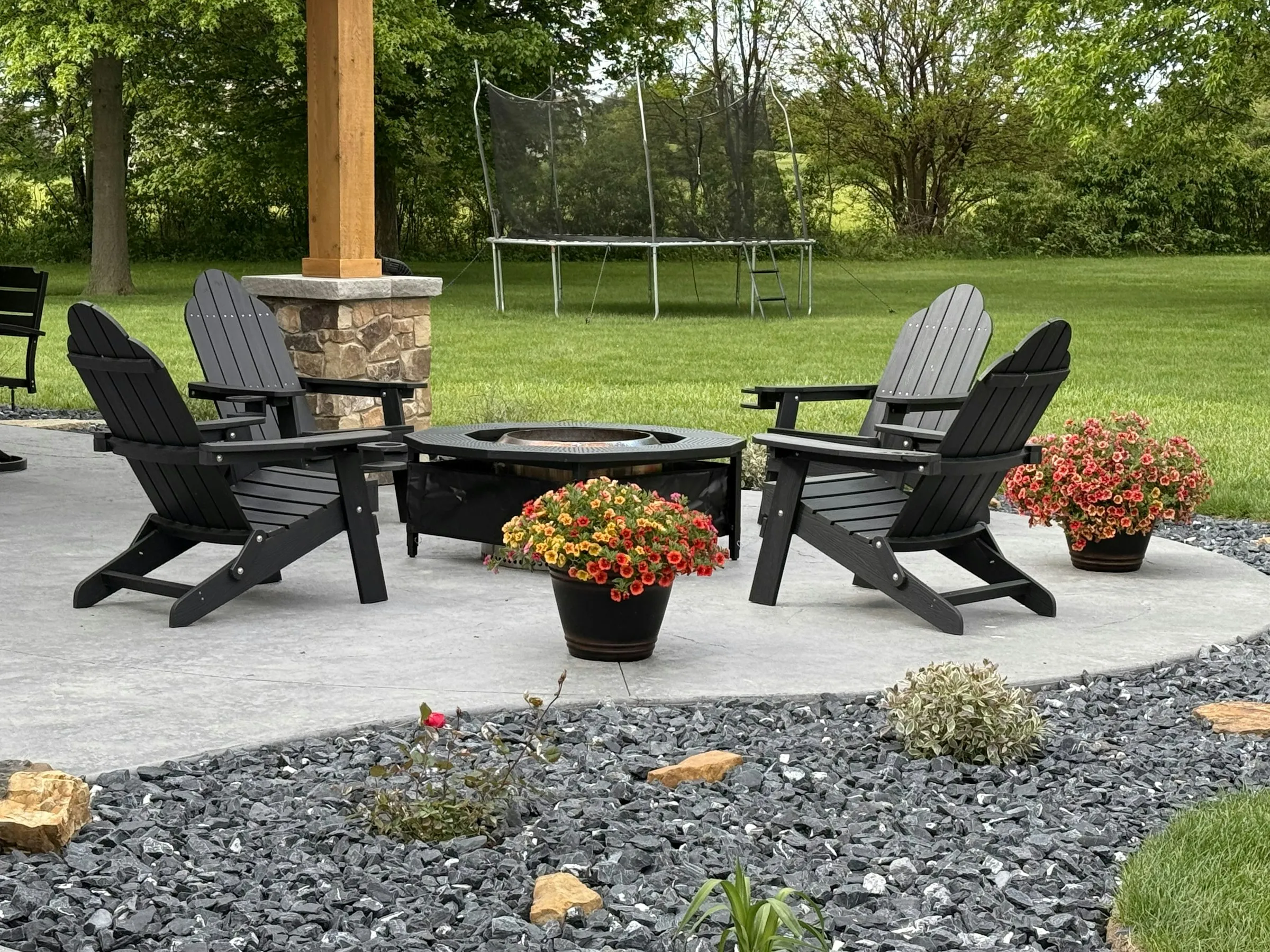 A finished concrete patio with four black adirondack chairs around a fire pit, edged with dark stone and flowers